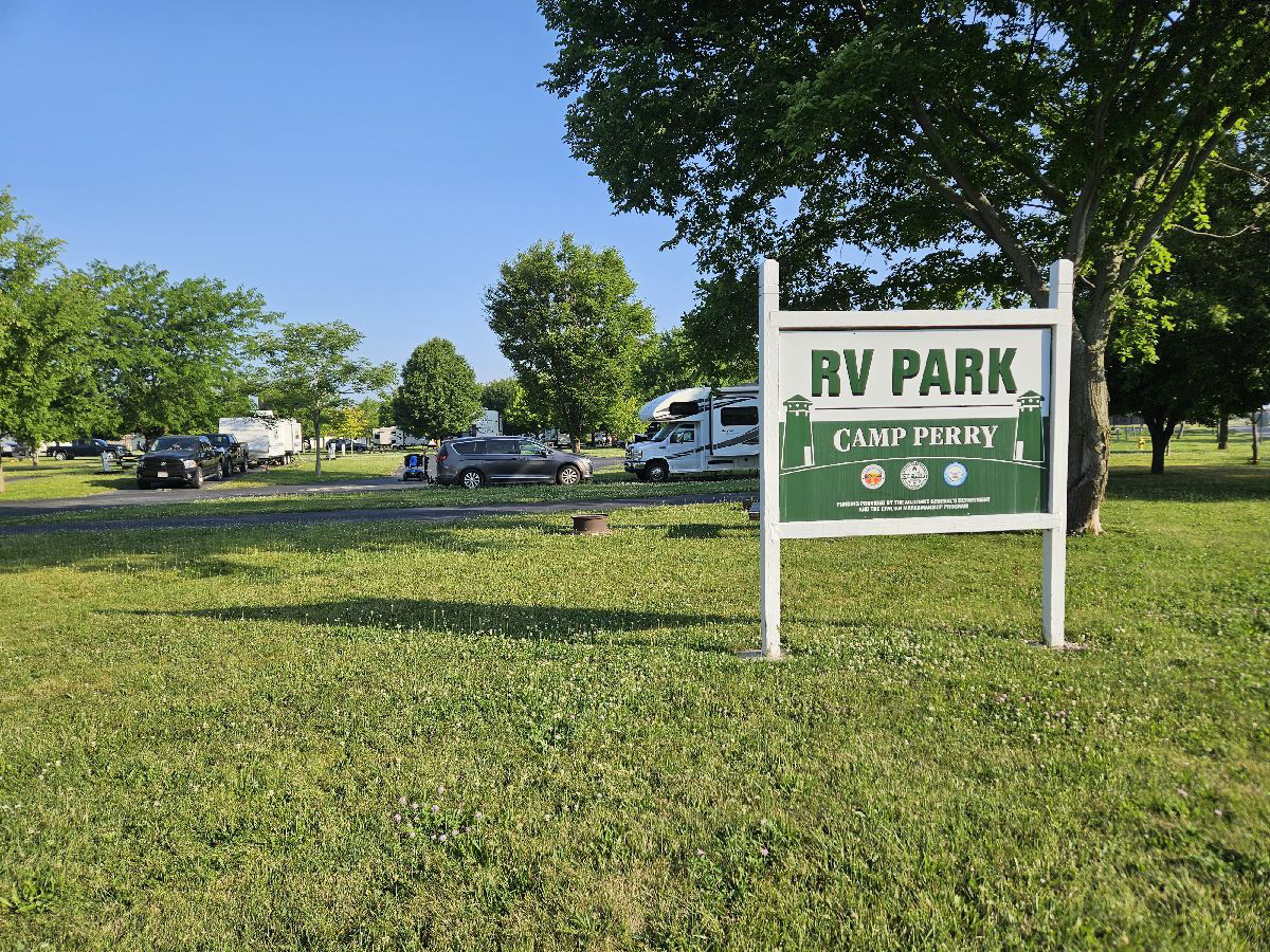 A green Camp Perry RV Park sign with rigs parked in the loop behind it